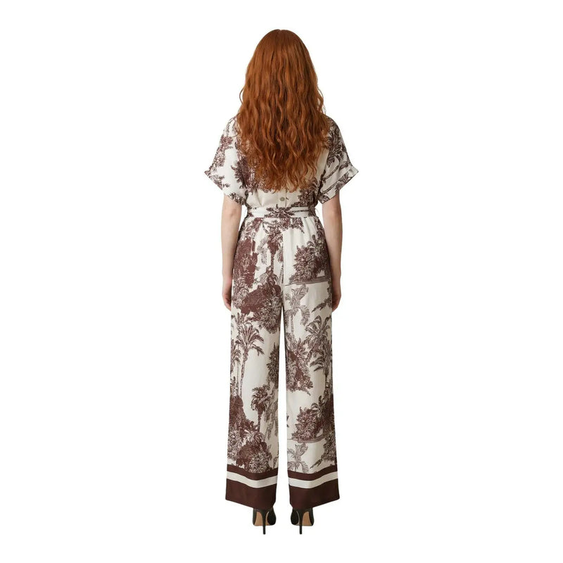 Eine Frau trägt einen beigefarbenen Artigli-Jumpsuit aus Viskose mit braunem Pflanzenmuster, flatternden Ärmeln und weiten Beinen.