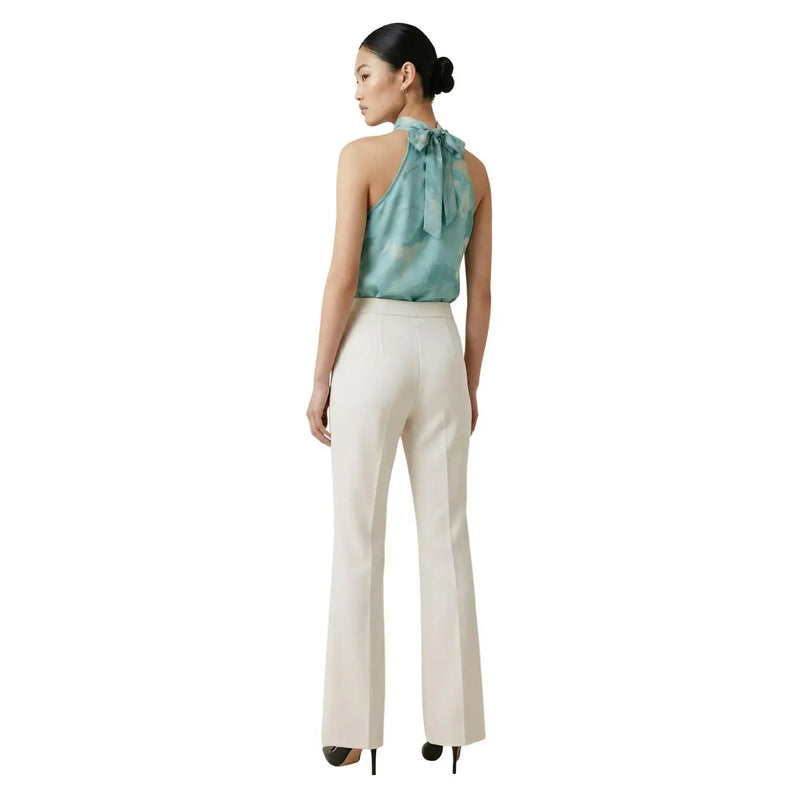 Eine Frau trägt eine weiße Artigli-Damenhose mit Reißverschlusstaschen vorne in Creme und ein türkisfarbenes Seiden-Neckholder-Top. top mit einer Schleife am Rücken