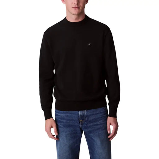 Männliches Model trägt einen schwarzen Rundhalspullover, ein graues Langarmshirt und eine schwarze Hose.