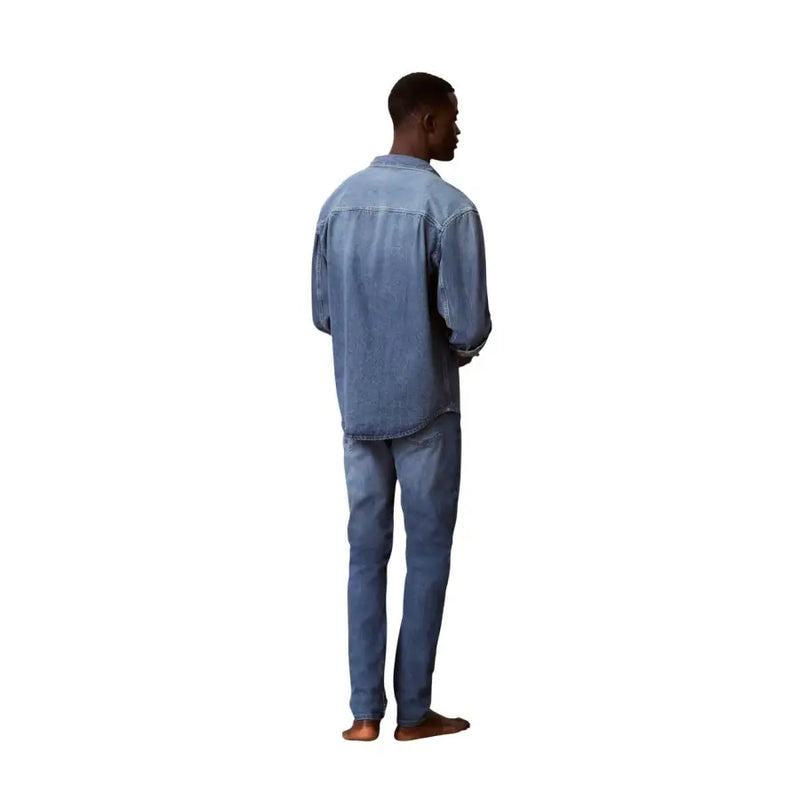 Ein Mann trägt eine schmal geschnittene, blaue Calvin Klein Herren-Jeans aus Baumwoll-Denim und ein dazu passendes hellblaues Jeanshemd.
