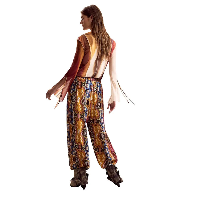 Eine Frau trägt eine senfgelbe Viskosehose von Desigual mit Gürtel, kombiniert mit einem farbenfrohen gemusterten Jumpsuit.