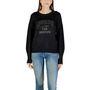 Woman wearing a black long-sleeved sweater with GUESS 1961 LOS ANGELES silver text and blue jeans