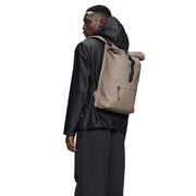 Beigefarbener Unisex-Rolltop-Rucksack von Rains mit schwarzen Akzenten und minimalistischem Design aus grauem Polyester. Maße: B 12 x H 48 x L 12 cm.