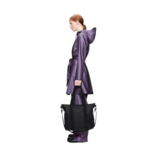 Eine elegante schwarze Mini-Tragetasche von Rains für Damen aus glänzendem Polyester mit stabilen Henkeln, gehalten von einem Model in einem violetten Regenmantel.