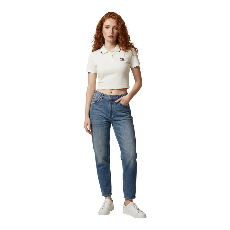 Eine Frau trägt ein cremefarbenes Tommy Hilfiger Polohemd mit schwarzem Besatz und blaue Replay Damenjeans, die die Vordertaschen freilegen.