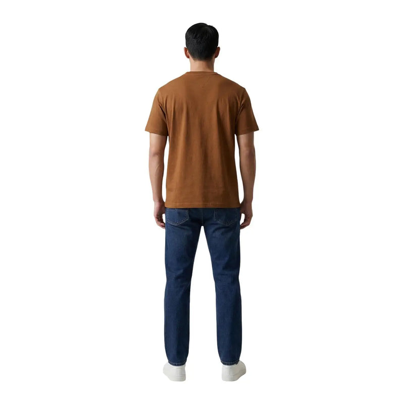 A man wearing a Tommy Hilfiger brown cotton round neck t shirt paired with dark blue jeans