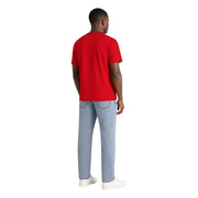Ein Mann trägt ein rotes, kurzärmeliges T-Shirt aus Bio-Baumwolle von Tommy Hilfiger und hellblaue Jeans.