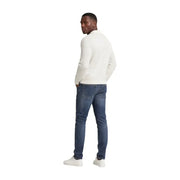 Ein Mann, der eine Tommy Hilfiger Herren Scanton Slim-Jeans aus blauer Baumwolle und dazu einen cremefarbenen, langärmeligen Strickpullover trägt.