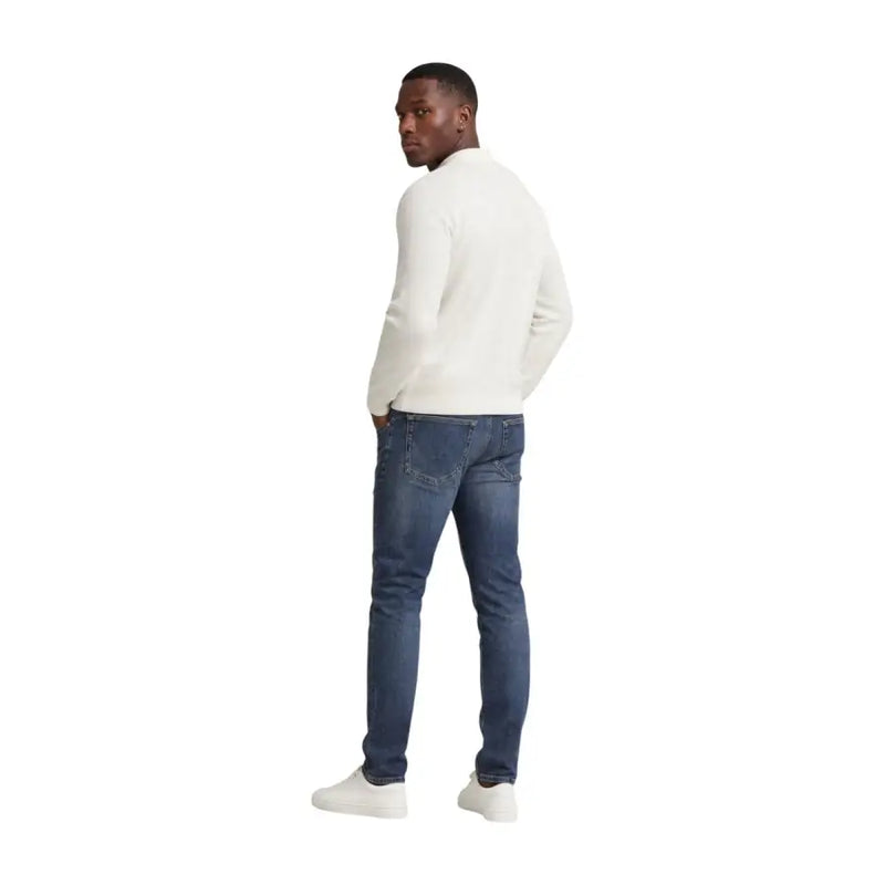 Ein Mann, der eine Tommy Hilfiger Herren Scanton Slim-Jeans aus blauer Baumwolle und dazu einen cremefarbenen, langärmeligen Strickpullover trägt.