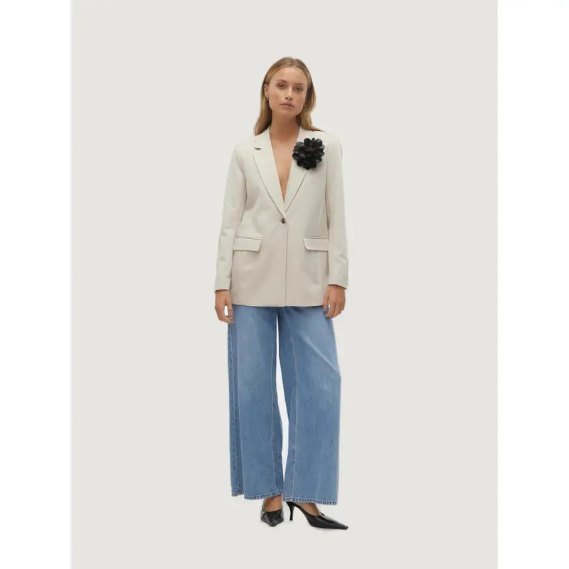 A woman wearing a vero moda beige blazer with button, front pockets, and a black floral brooch on the left lapel