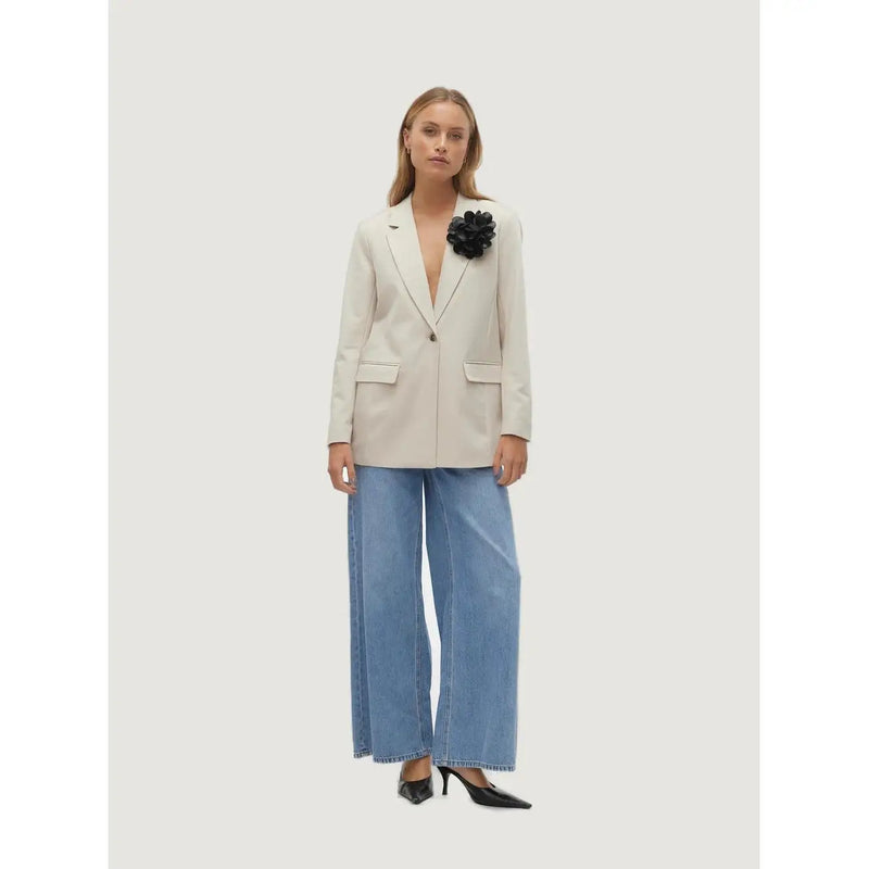 A woman wearing a vero moda beige blazer with button, front pockets, and a black floral brooch on the left lapel
